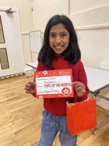 Little girl with long dark hair wearing a red top and jeans, carrying a red paper bag and holding a certificate that says Little Voices Pupil of the Month
