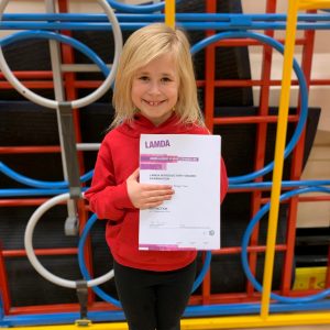 Young girl with blonde hair wearing a red Little Voices hoodie. She's standing in front of a multi-coloured climbing frame in a school gym and holding up a LAMDA certificate.