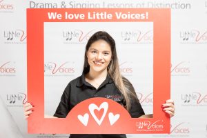 Ashlea Bateup Little Voices South West London franchisee. Woman with long brown hair and black t-shirt holding red photo frame with the words We Love Little Voices in white