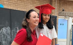 Young asian girl wearing a red mortar board on her head. Her Little Voices tutors is kneeling beside her and they are both smiling. This photo was taken during the cost of living crisis 2023.
