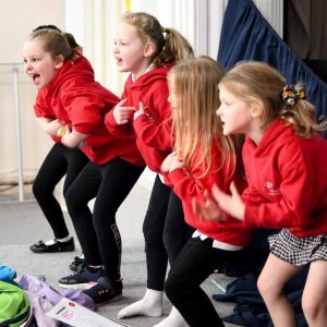 Four children in a line on a stage. They're wearing red Little Voices hoodies and are acting out a scene from a play where they're pointing wildly at themselves.