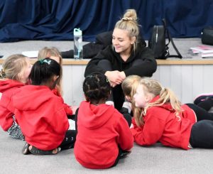 A group of young children wearing red Little Voices hoodies sitting in a circle with a Little Voices tutor at the front.