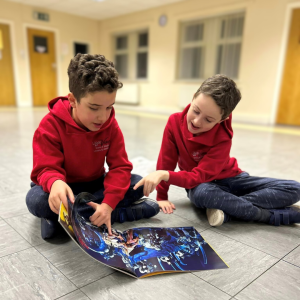Two young boys sitting cross legged on the floor. They're wearing red Little Voices hoodies and are looking at a book together, demonstrating teamwork skills.