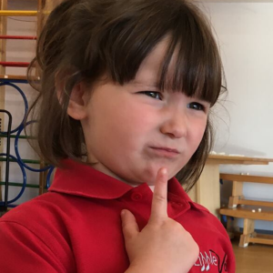 Young girl wearing a red Little Voices polo shirt. She has her finger on her chin and looks like she's thinking about something.